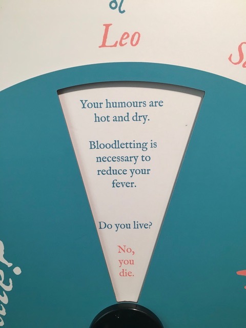 Photo depicts a section of an interactive label where the text is visible through a keyhole window on a turning dial. It says "Your humours are hot and dry. Bloodletting is necessary to reduce your fever. Do you live?" and in red color text, it says "No, you die."